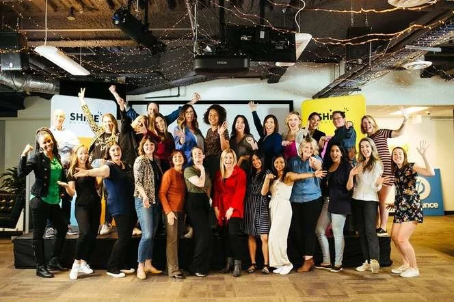 A large group of Shine Bootcamp participants and coaches posing together in a brick-walled venue