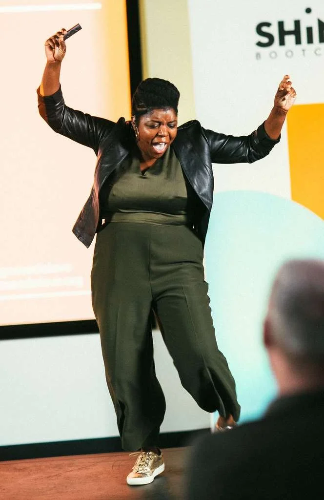 A Shine speaker mid-stride on stage, microphone in hand and arm raised