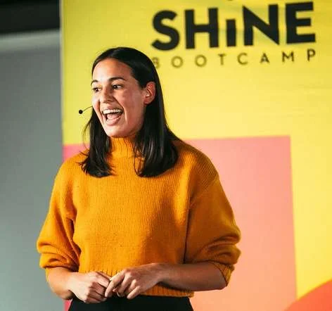 A smiling Shine speaker in a mustard sweater in front of the Shine Bootcamp logo