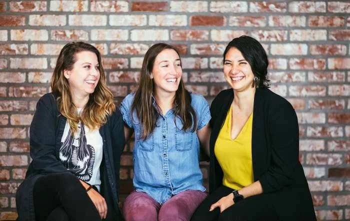 Three Shine participants laughing together on a sofa in front of a brick wall
